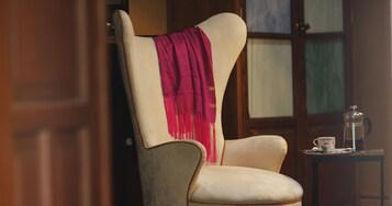 Una silla con un pañuelo rosa, una mesa con una tetera y una taza, y una puerta de madera al fondo.
