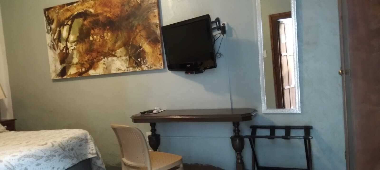 Una habitación con cama, un escritorio con silla, un televisor montado en la pared y un cuadro.
