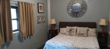Un dormitorio con una cama, dos mesitas de noche con lámparas, un espejo en la pared y dos cuadros enmarcados.