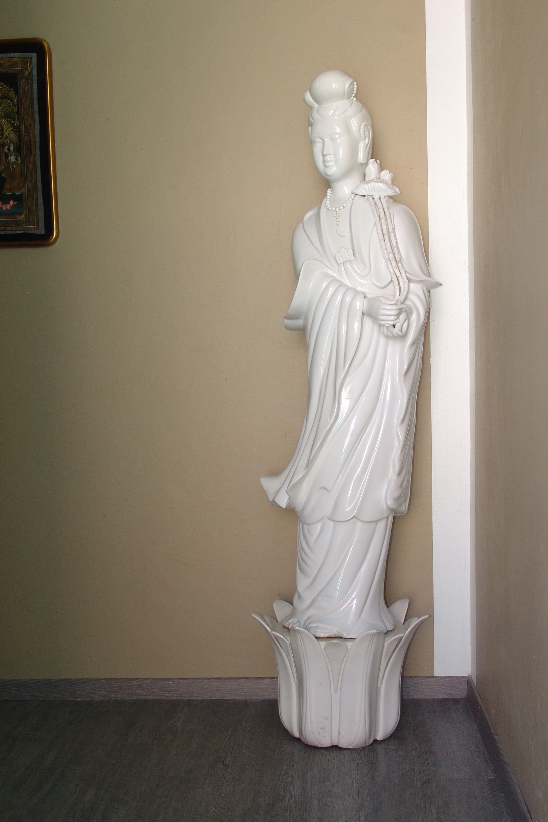 Une statue blanche représentant une figure vêtue d’une tenue traditionnelle, placée devant un mur beige et sur un plancher en bois foncé.