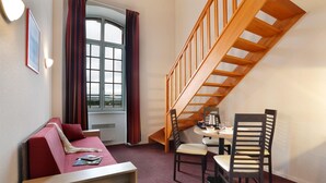 Una habitación con escalera de madera, un sofá rojo, una pequeña mesa de comedor con dos sillas y una ventana con cortinas.