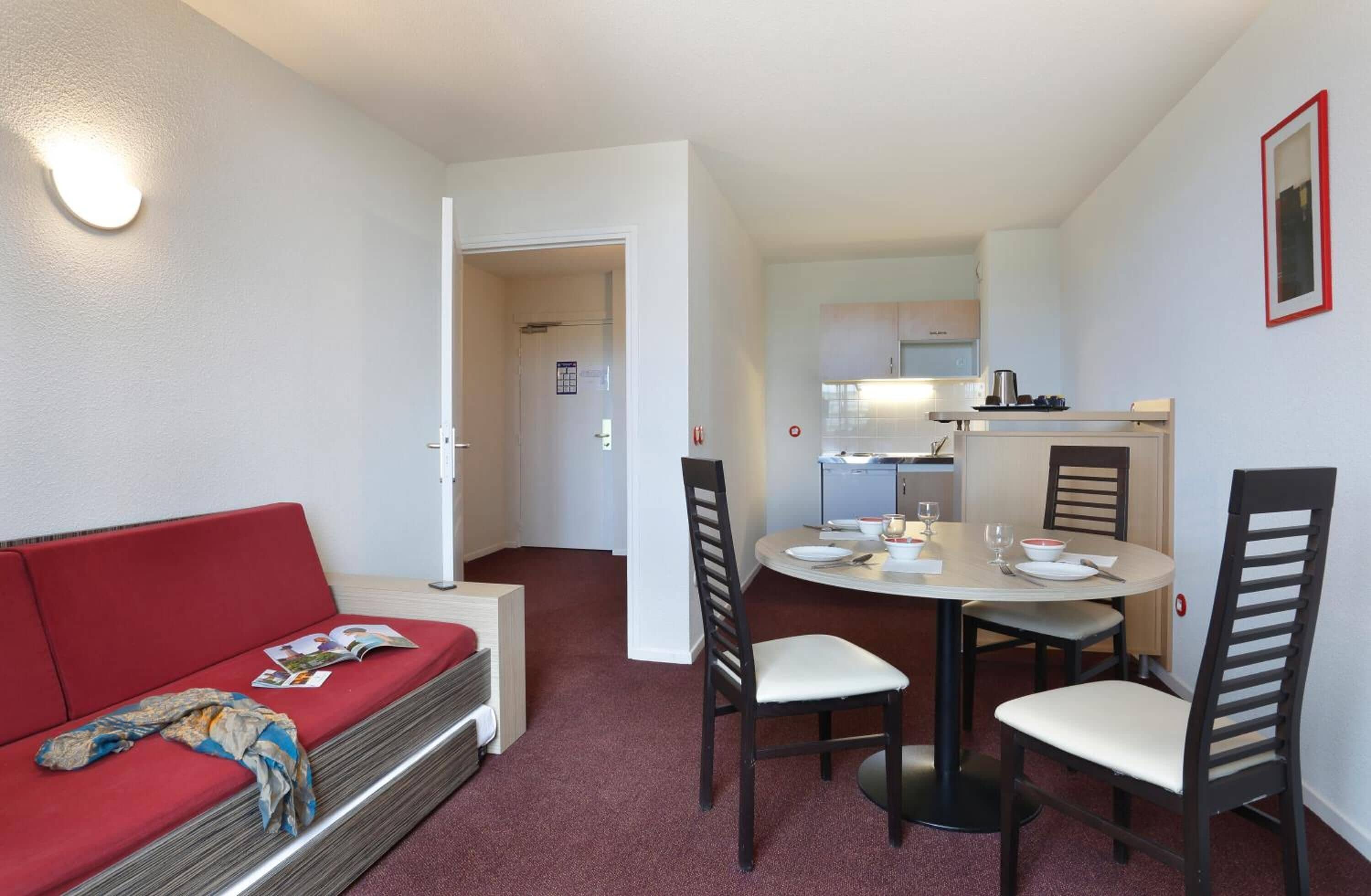 A compact living space with a red sofa, a dining table with four chairs, and a kitchenette in the background.