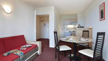 A compact living space with a red sofa, a dining table with four chairs, and a kitchenette in the background.