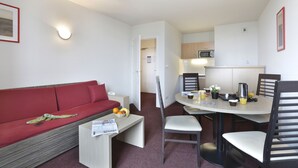 Un espacio habitable compacto con un sofá rojo, una pequeña mesa de comedor con sillas, una kitchenette y una pared blanca con una lámpara.