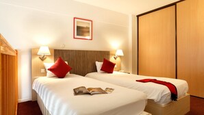 Habitación de hotel con dos camas, cada una con ropa blanca y almohadas rojas, cabeceros de madera, lámparas de noche y un armario de madera.