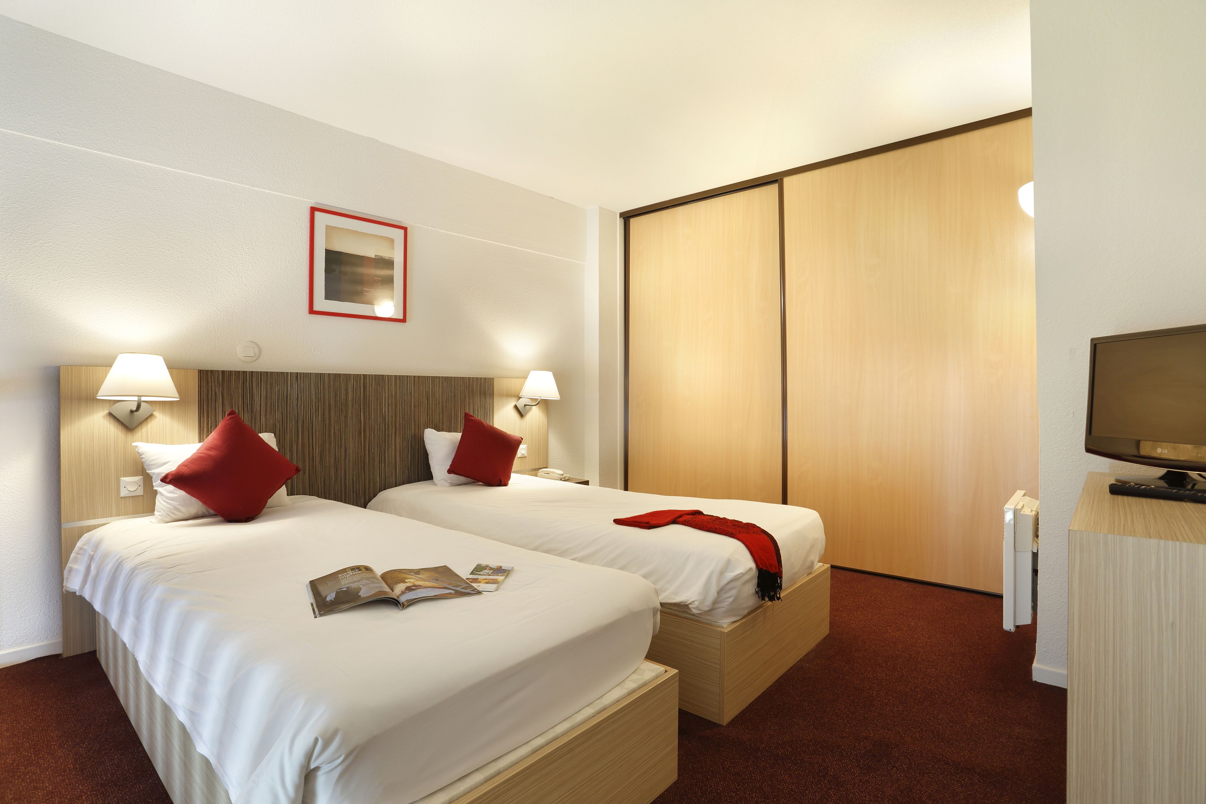 Ein Hotelzimmer mit zwei Betten, jeweils mit weißer Bettwäsche und roten Kissen, einem hölzernen Kopfteil, Nachttischlampen, einem Holzschrank und einem Fernseher auf einem Holzschrank.