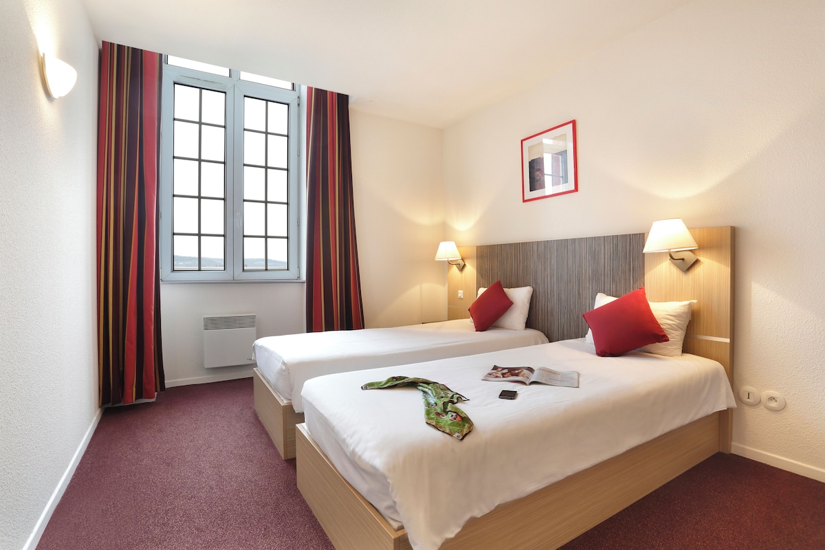 Habitación de hotel con dos camas, una ventana con cortinas, un espejo en la pared y dos lámparas de pared.