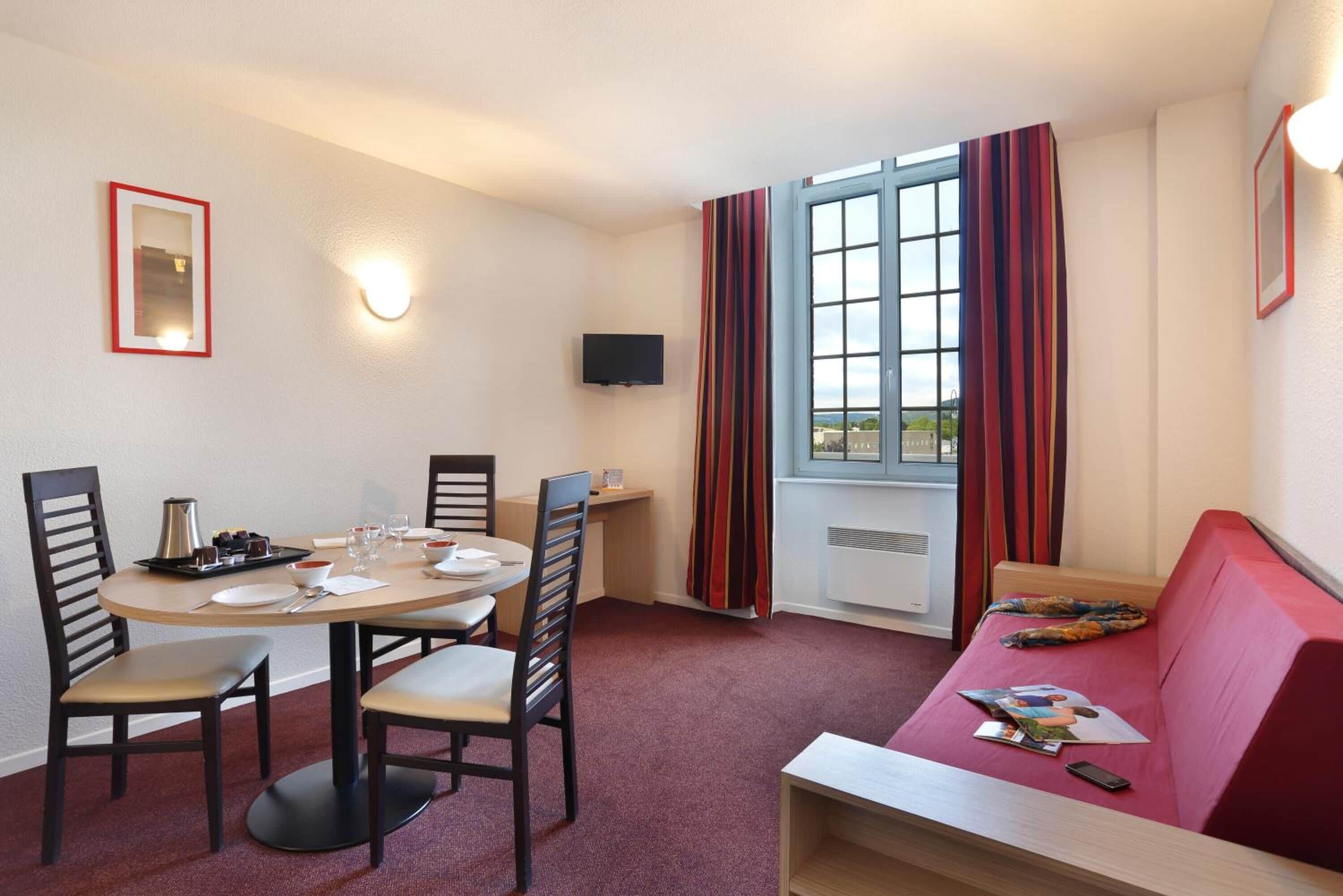 A hotel room with a dining table, chairs, a sofa, a TV, and a window with curtains.