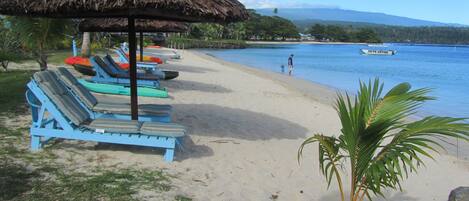 Una playa con sillas de descanso y una sombrilla de paja, una pequeña palmera y un barco llamado 'DIVE SAVAUI' en el agua.
