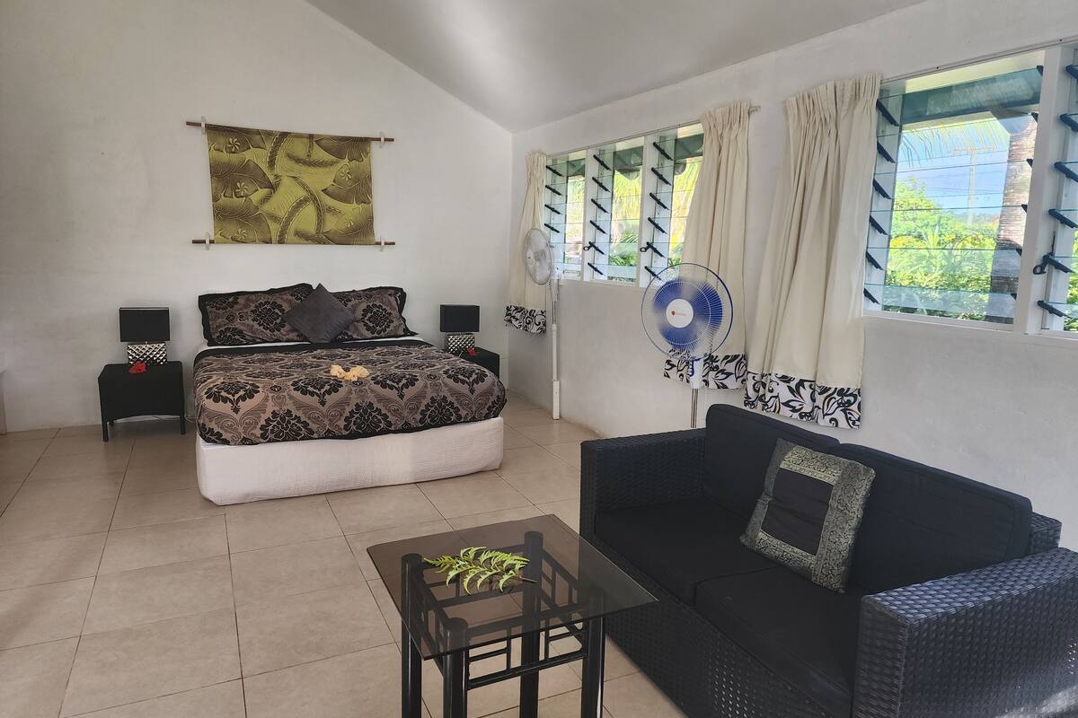 A bedroom with a bed, bedside tables, a fan, a sofa, and a window with curtains.