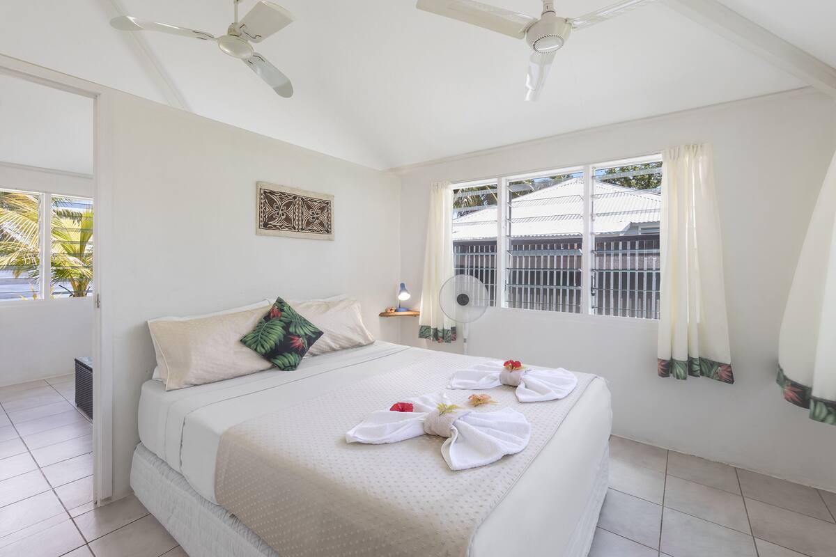 A hotel room with a bed, ceiling fans, a window with curtains, and a wall-mounted fan.