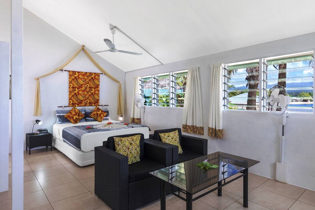 A bedroom with a bed, two black sofas, a glass table, a ceiling fan, and a window with a view of palm trees and mountains.