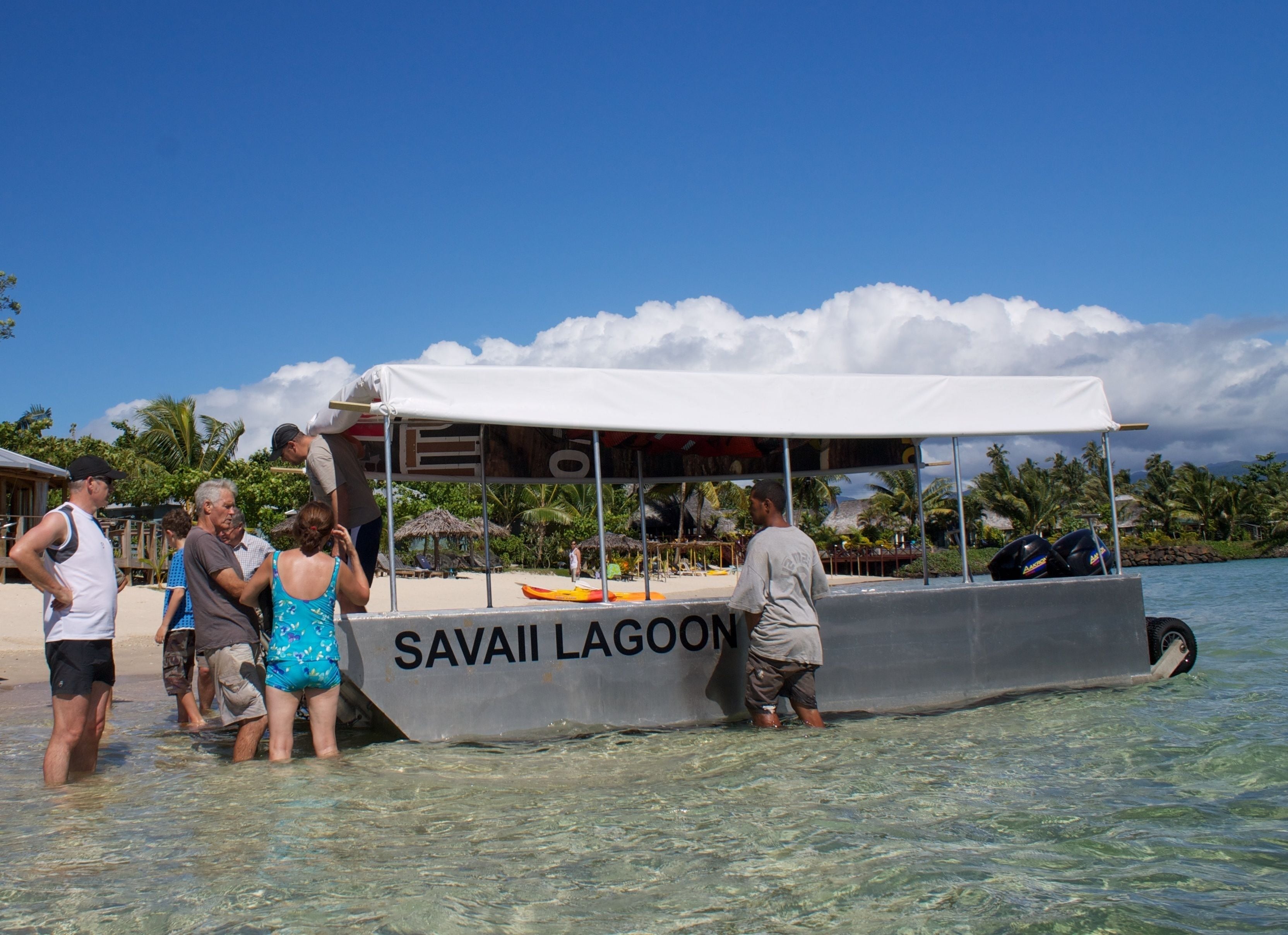 Un bote llamado "Savai'i Lagoon" con cubierta techada y algunas personas de pie en la proa.
