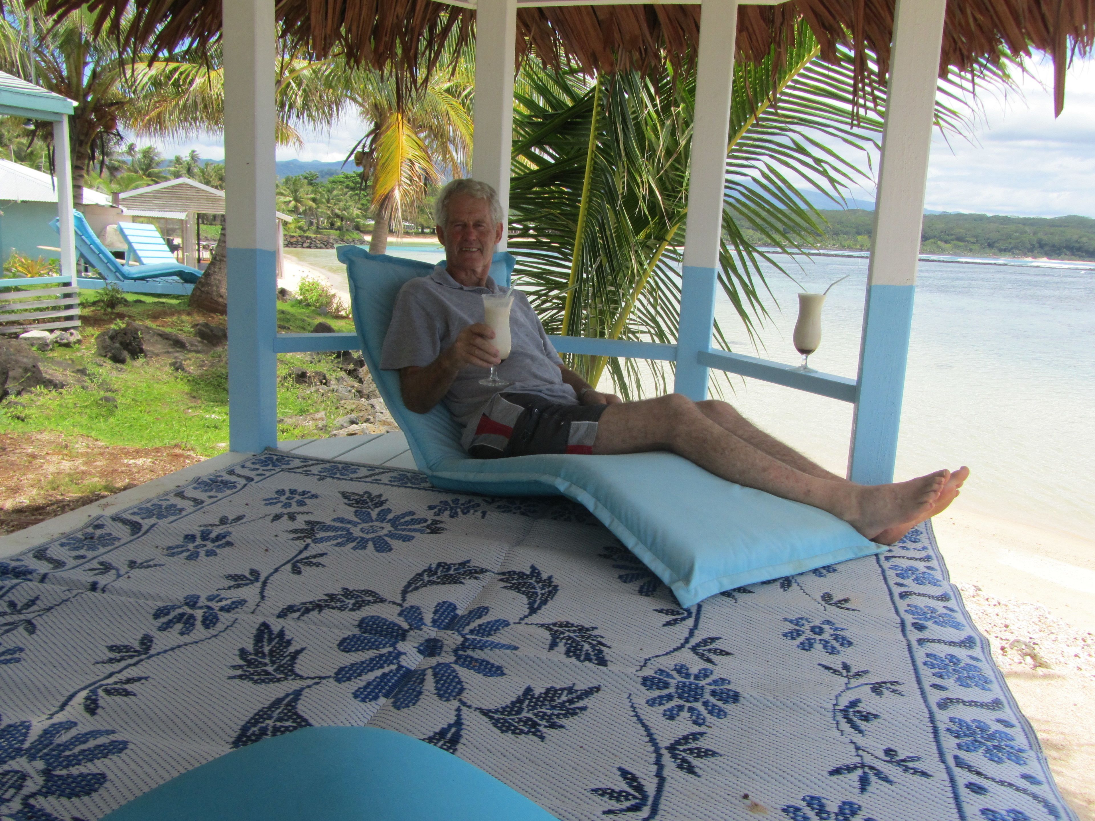 Un homme se détend sur un fauteuil bleu, avec un coussin à motifs floraux, et tient une boisson à la main.