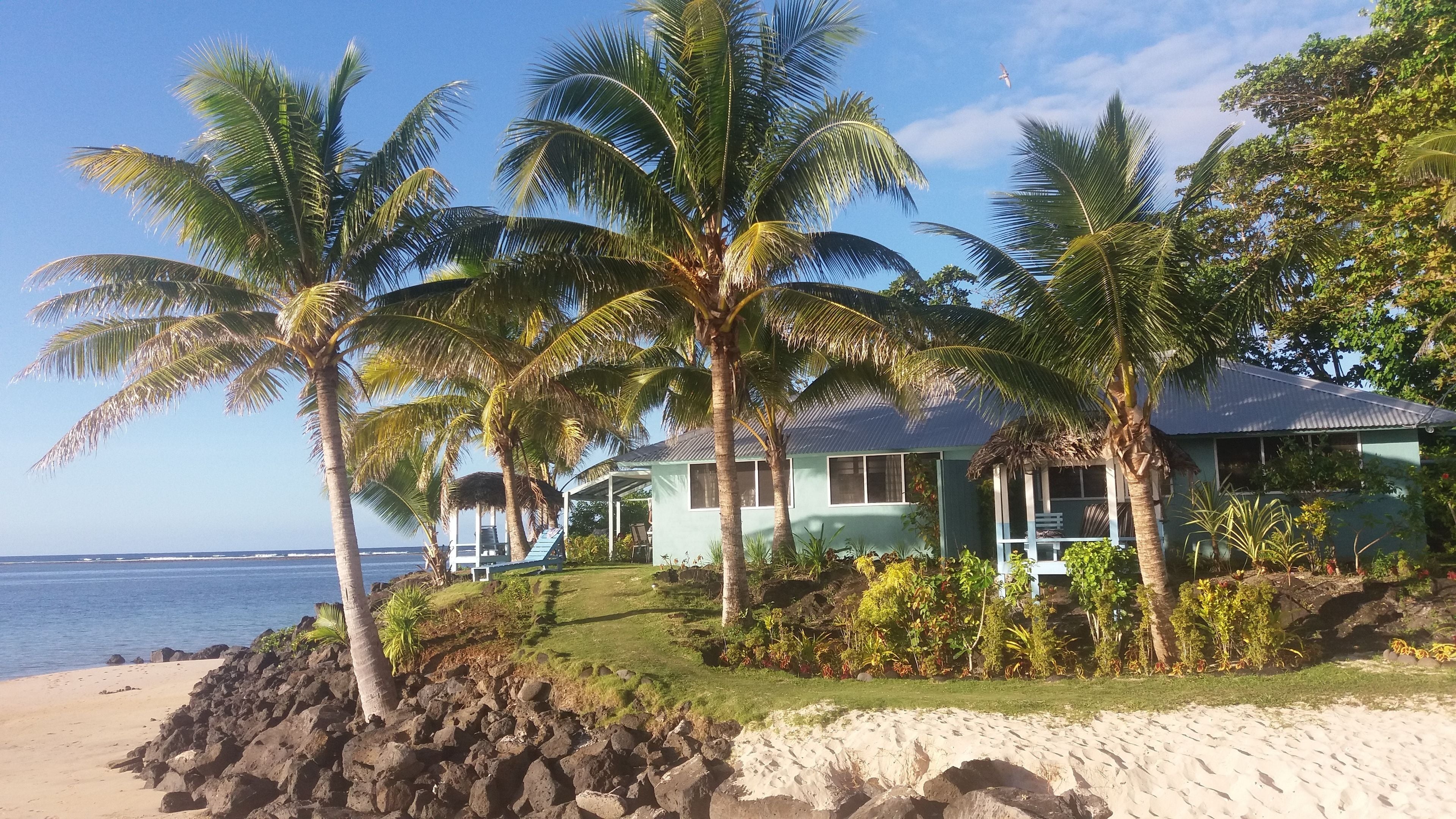 A beachfront property with a house surrounded by palm trees, a rocky shoreline, and a view of the ocean.