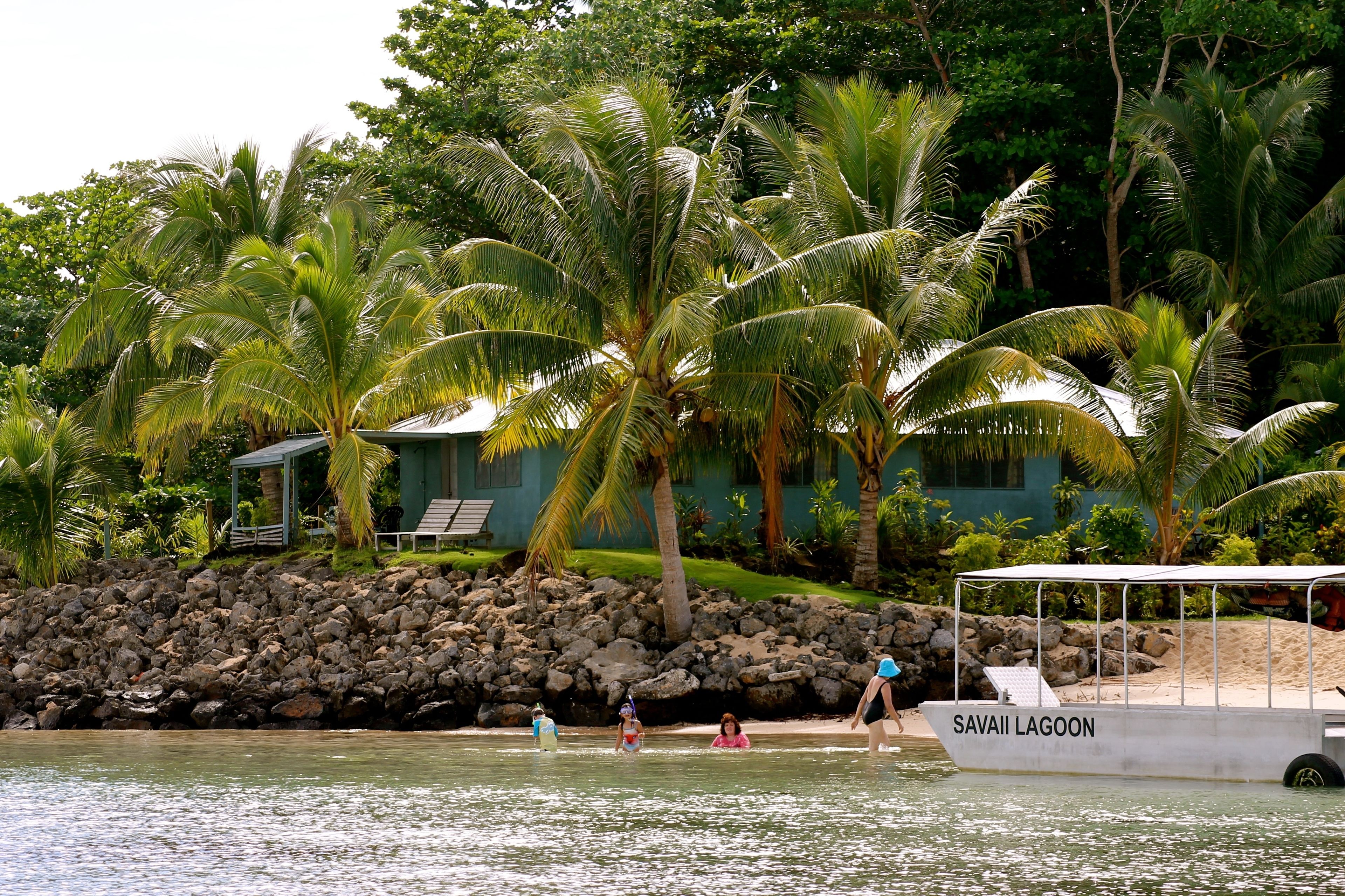 A boat named "Savai'i Lagoon" docked near a rocky shore with palm trees and a building in the background.