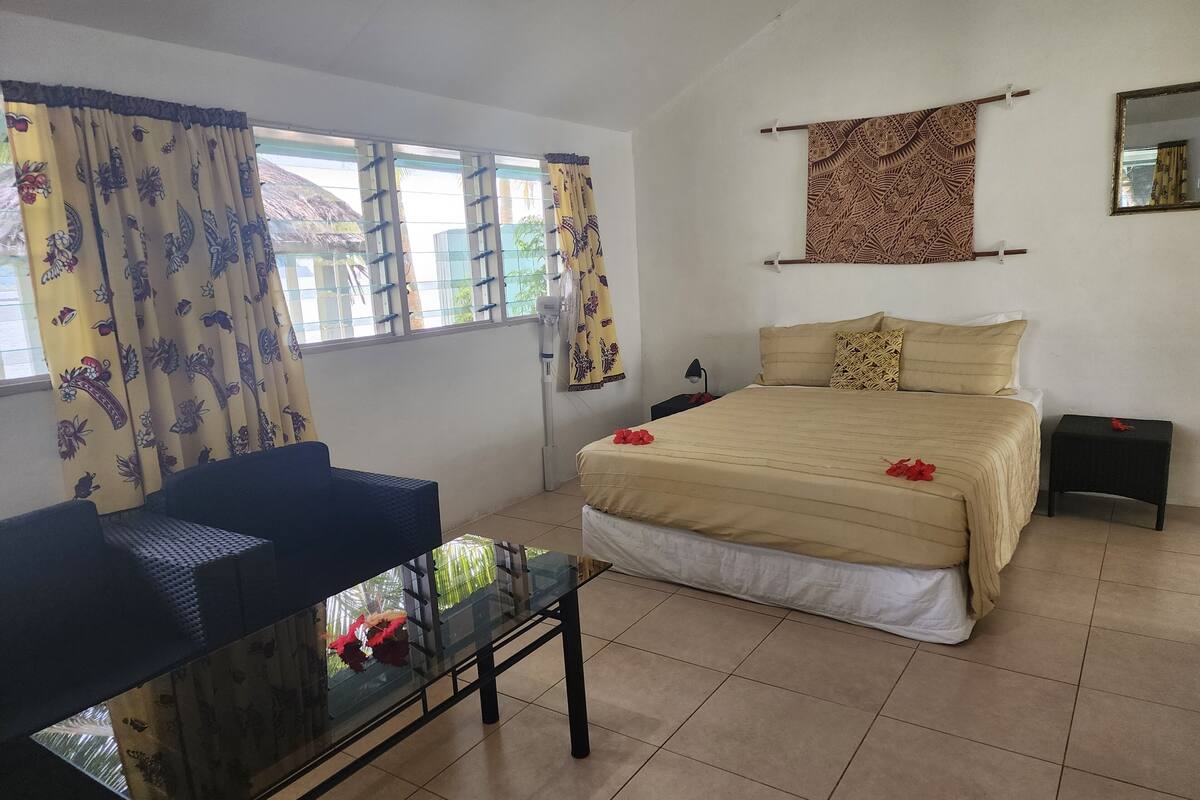 A bedroom with a bed, a chair, a small table, a nightstand, a window with curtains, and a wall-mounted mirror.