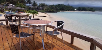 A wooden deck with metal chairs and a table overlooking a beach with clear water and palm trees.