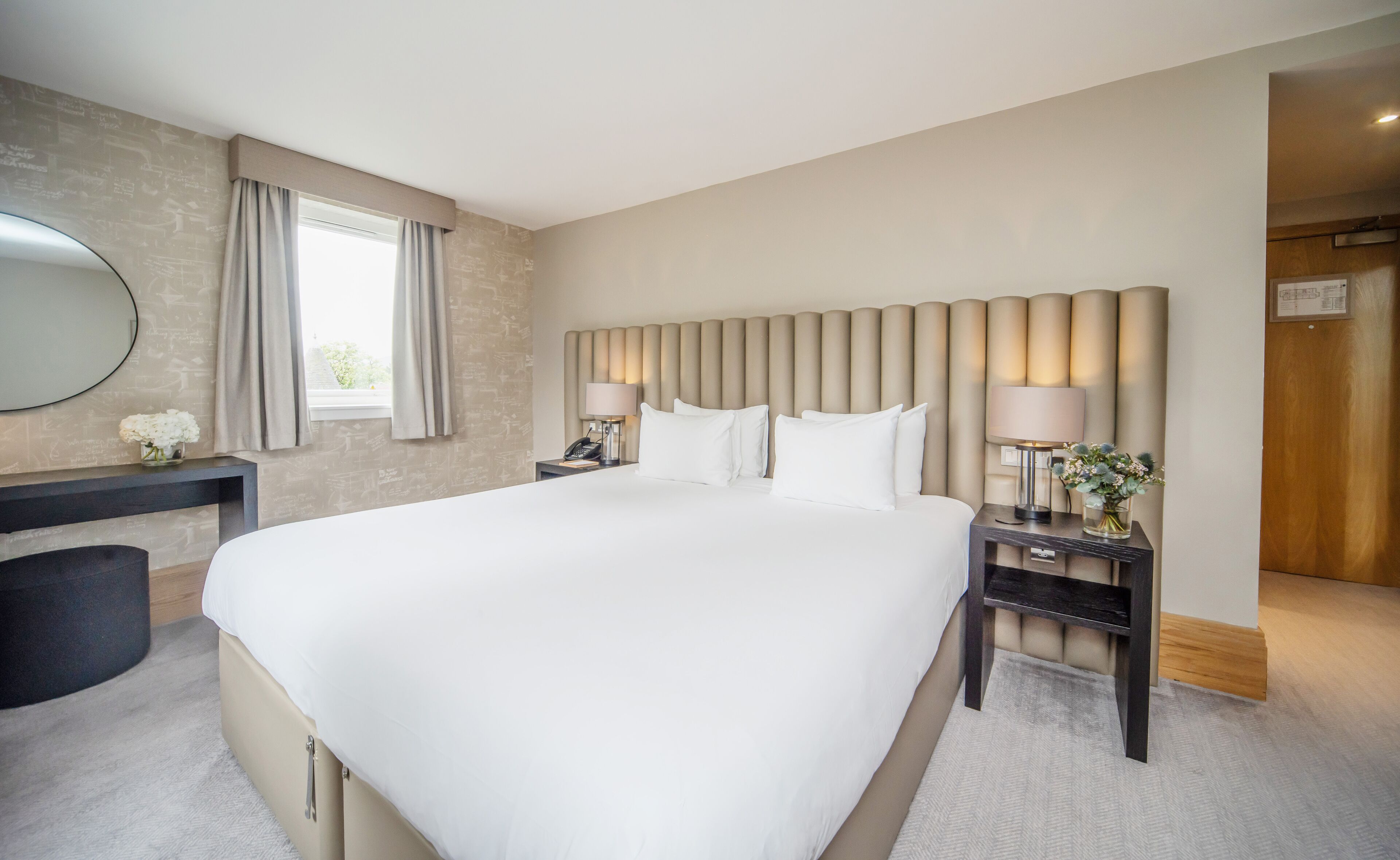 A hotel room with a large bed, a bedside table with a lamp, a desk with a mirror, and a window with curtains.