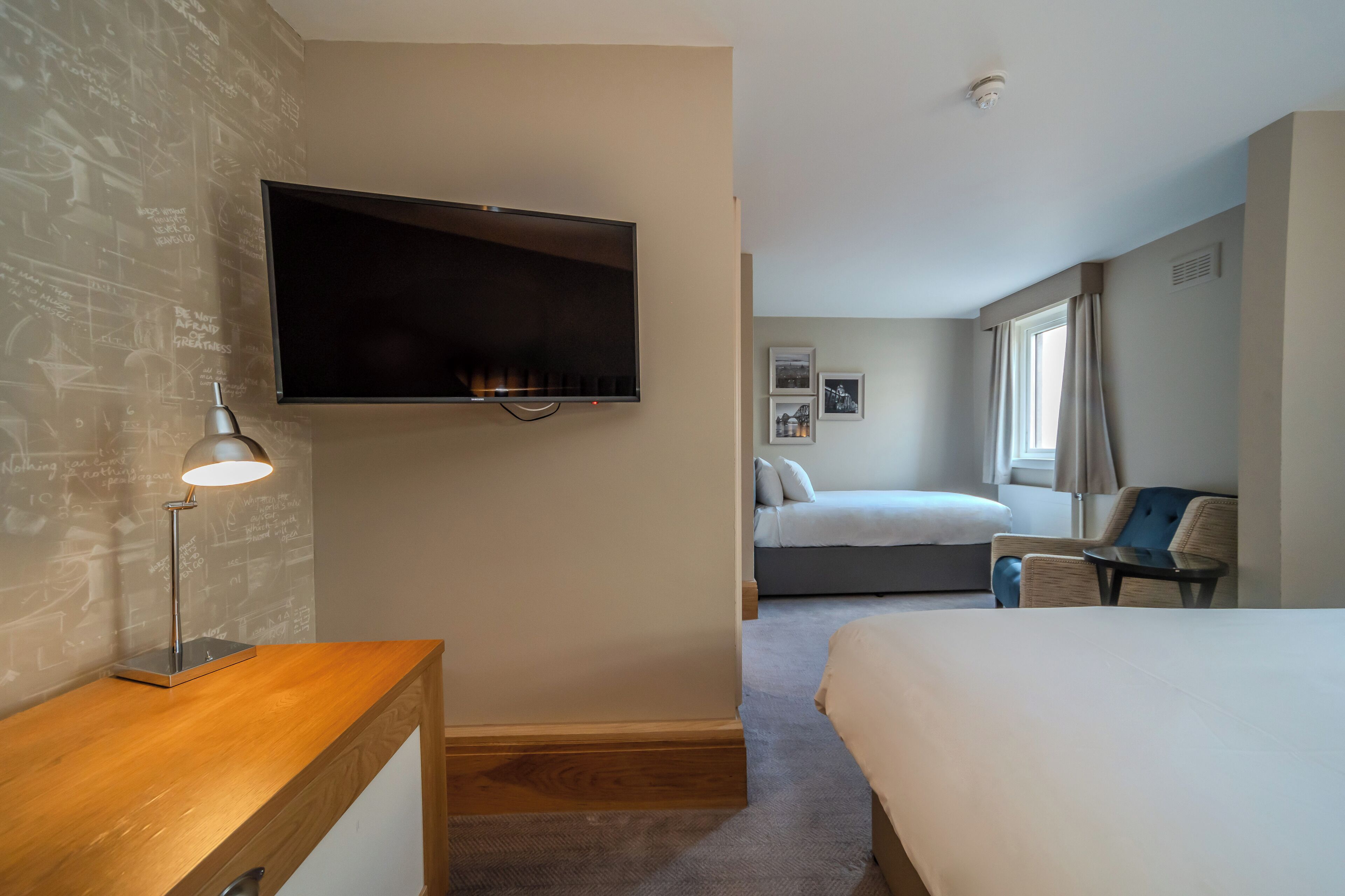 Een hotelkamer met een bed, een bureau met lamp, een aan de muur bevestigde televisie, een raam met gordijnen en een klein tafeltje met stoel.