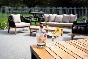 A bottle of Pickering's gin and a cocktail on a wooden table in an outdoor patio area with a sofa and chairs.