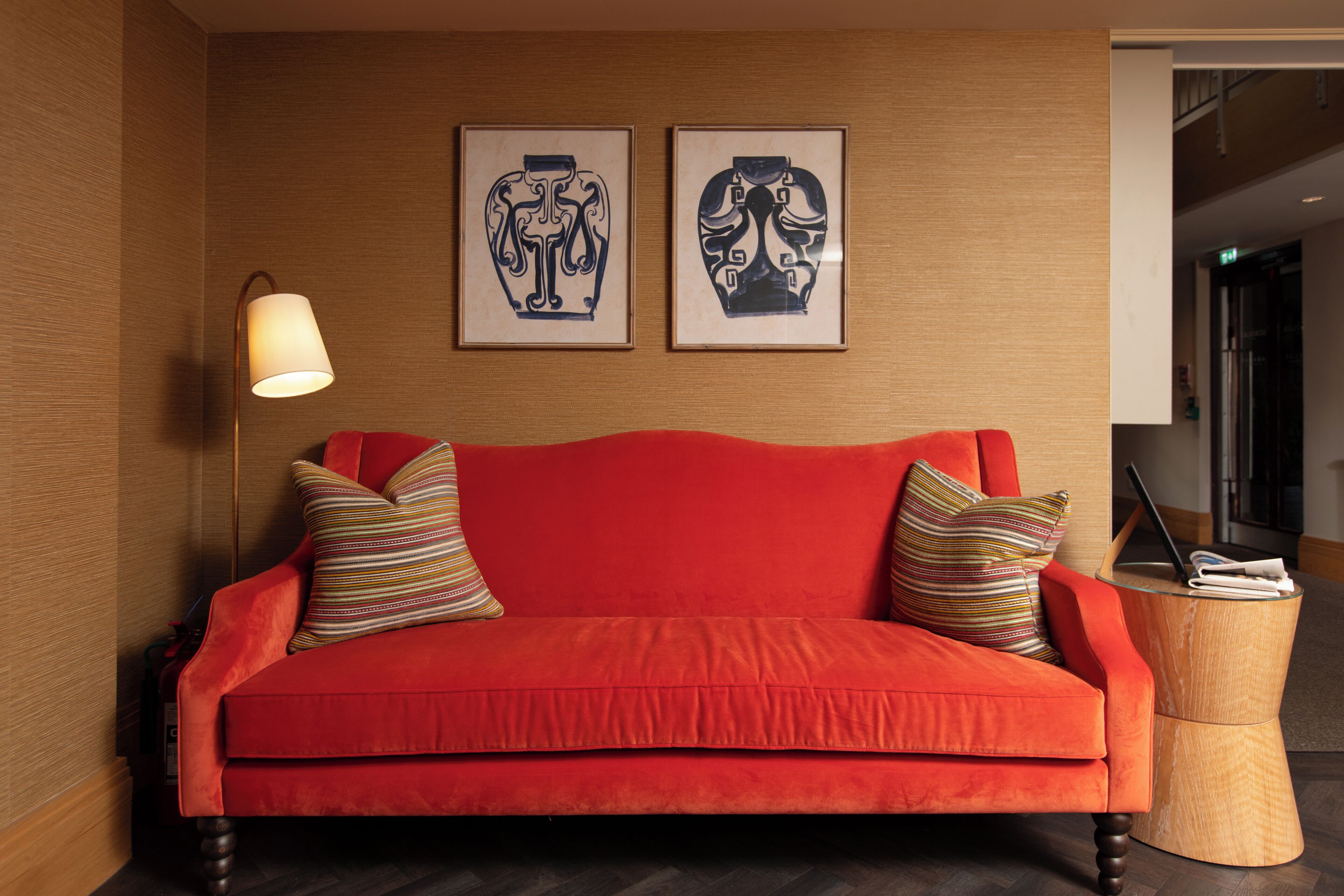 Een rode bank met gestreepte kussens, een houten bijzettafeltje met een lamp, en twee kunstwerken in lijsten aan de muur.