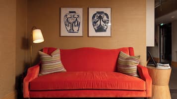 Een rode bank met gestreepte kussens, een houten bijzettafeltje met een lamp, en twee kunstwerken in lijsten aan de muur.