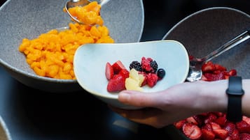 A person holding a bowl with mixed fruits, with another bowl containing mangoes in the background.