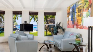 A modern living room with two grey sofas, a coffee table, and a TV. Large windows offer views of a beach and palm trees. - Azul del Mar (Key Largo)