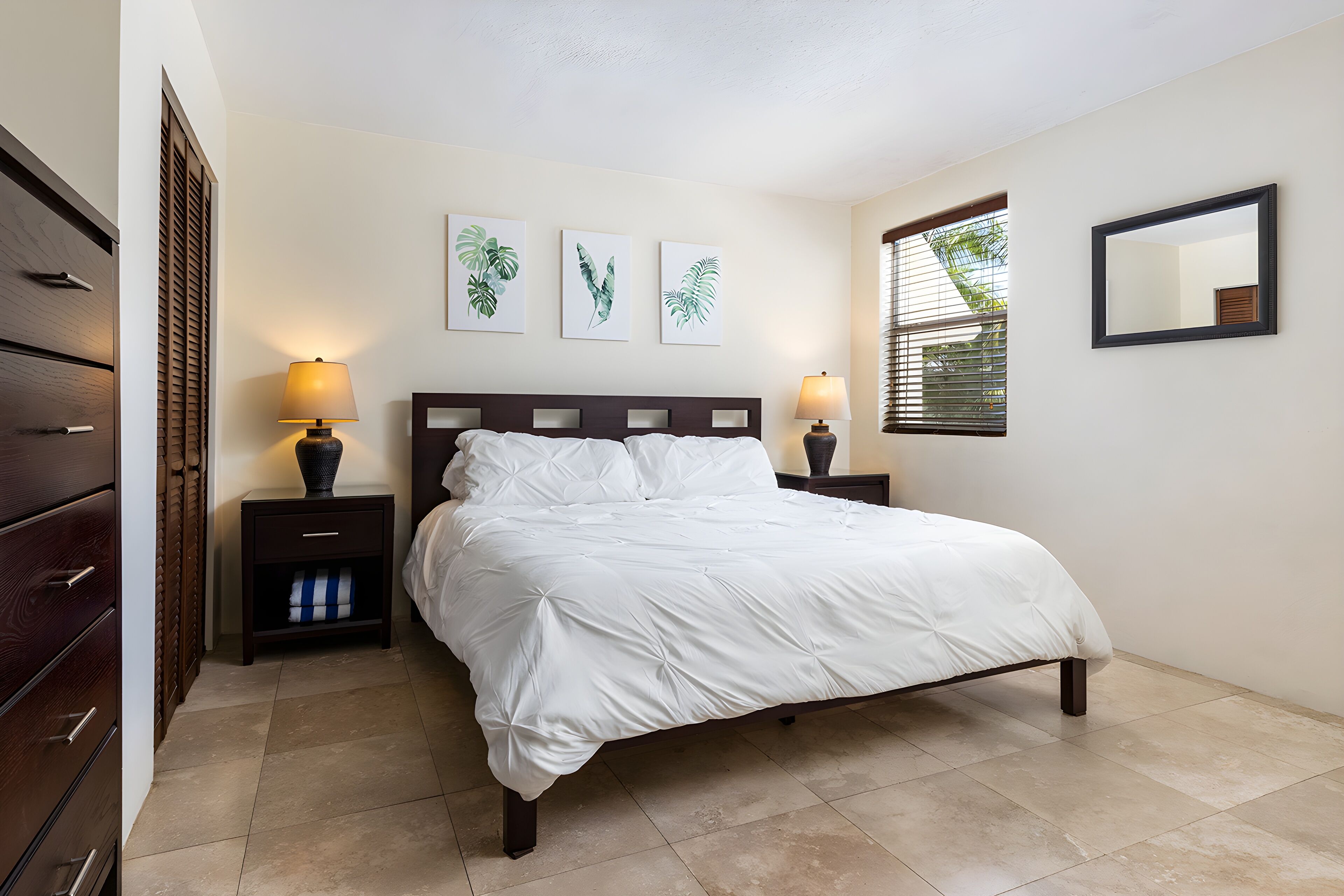 A bedroom with a bed, two bedside tables with lamps, a dresser, and three wall-mounted plant prints.