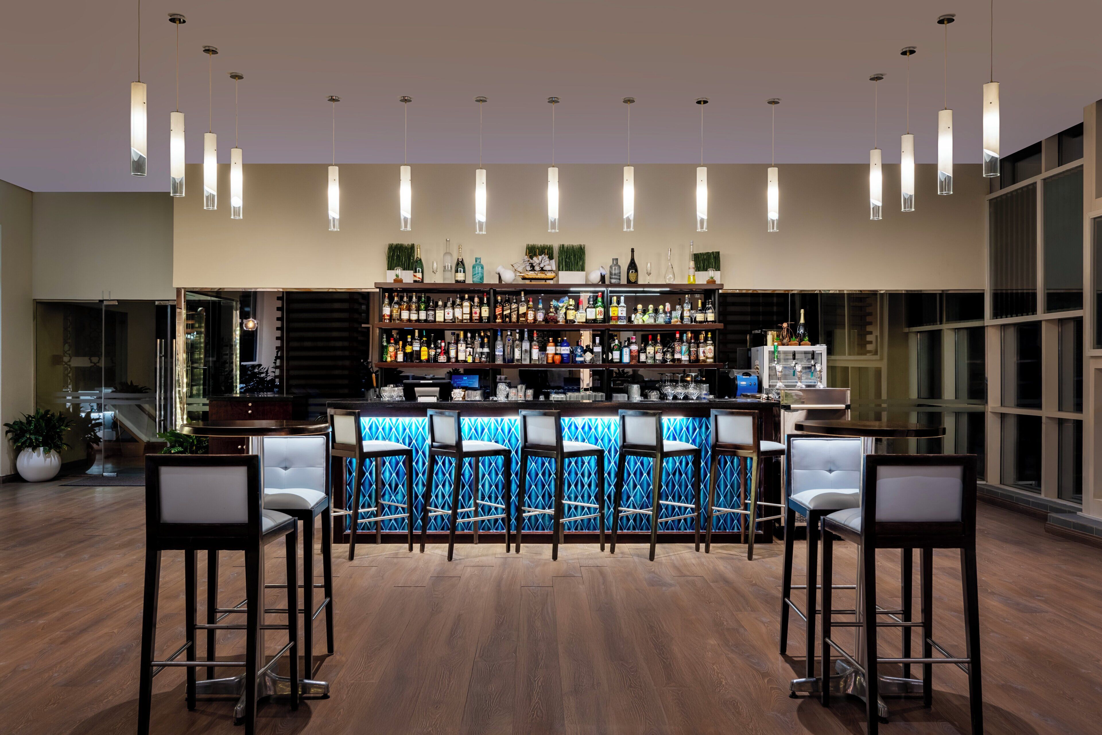 A bar area with a wooden floor, bar stools, and a well-stocked shelf with various bottles.