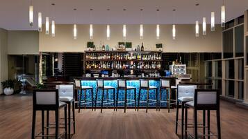 A bar area with a wooden floor, bar stools, and a well-stocked shelf with various bottles.
