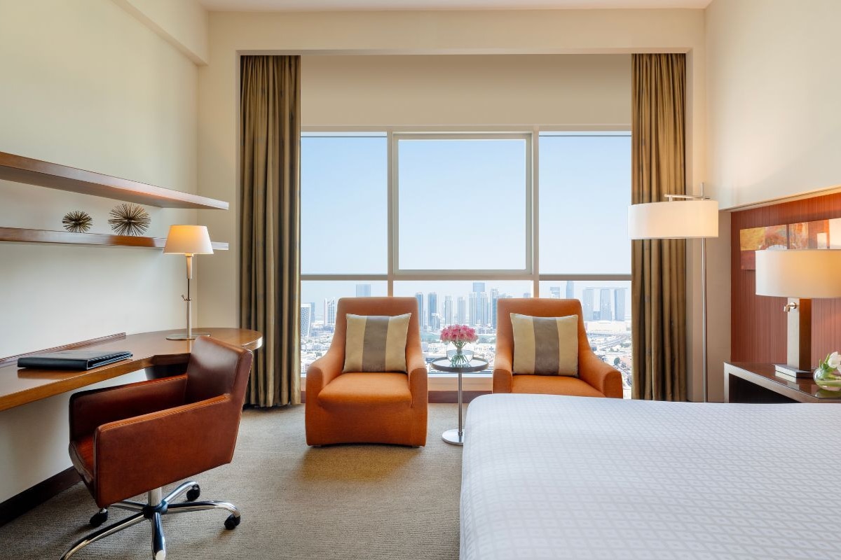 Habitación de hotel con escritorio, dos sillones, una cama, vistas a la ciudad y una pequeña mesa con flores.
