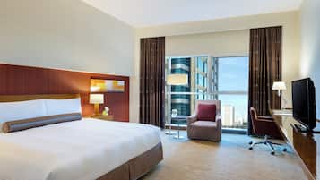 Une chambre d’hôtel avec un grand lit, un bureau avec une télévision, une chaise et une vue sur la ville grâce à de grandes fenêtres.