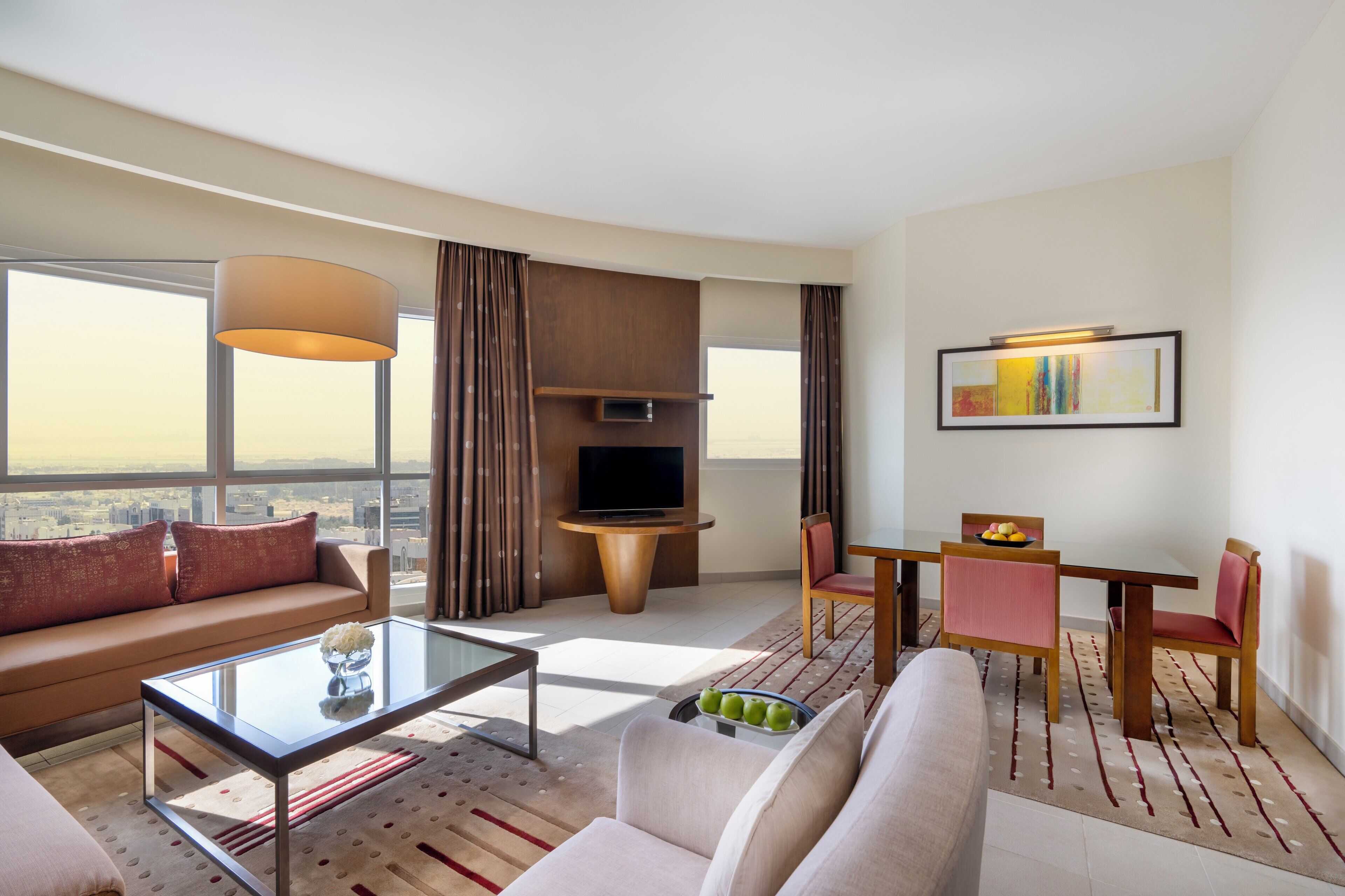 A modern hotel room with a sofa, a glass coffee table, a dining area with a table and chairs, a TV, and a large window offering a city view.