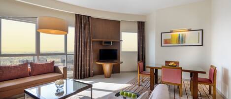 A modern hotel room with a sofa, a glass coffee table, a dining area with a table and chairs, a TV, and a large window offering a city view.