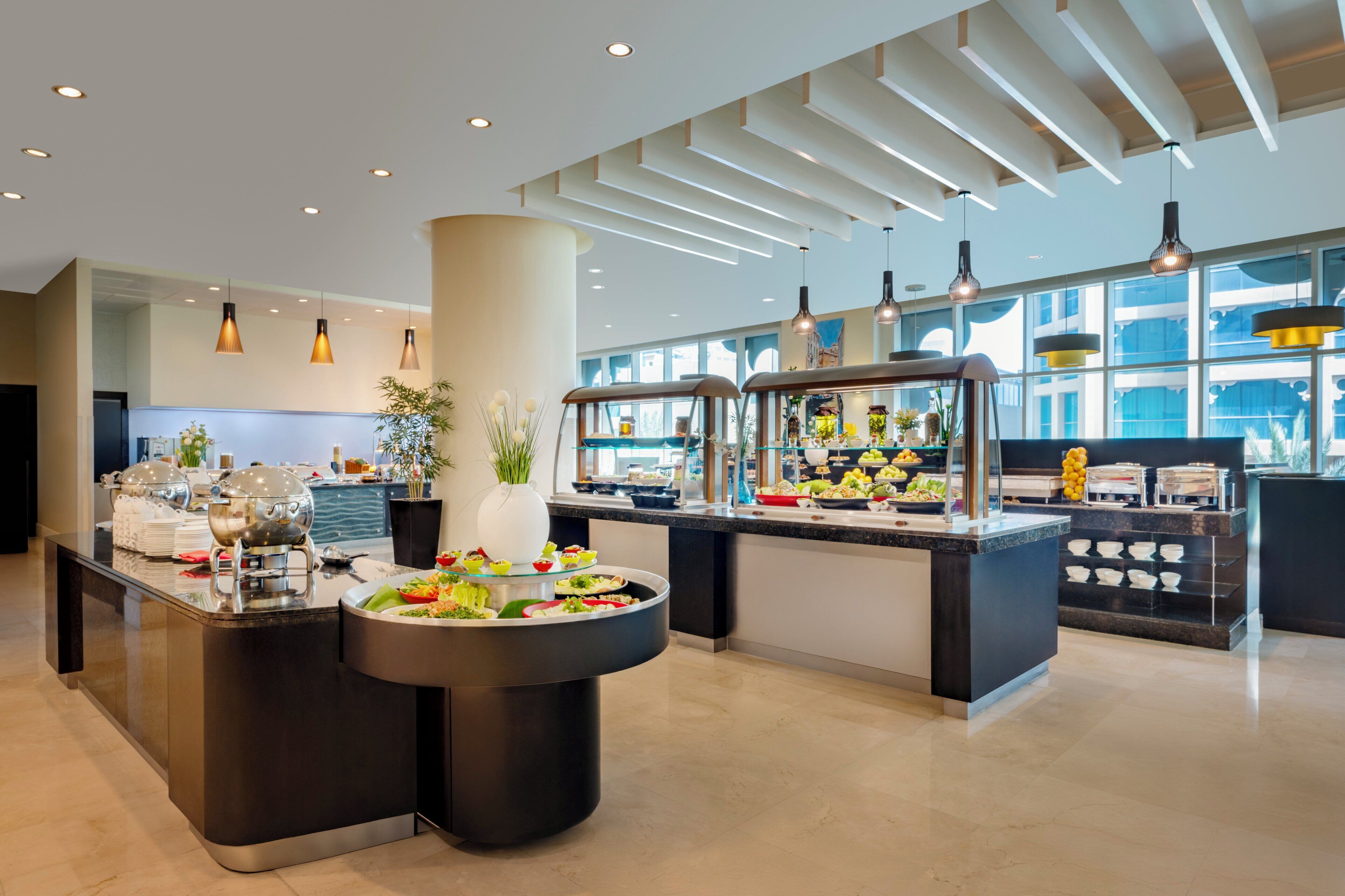 A modern hotel lobby with a buffet area featuring various food stations and a large central island with a decorative plant.
