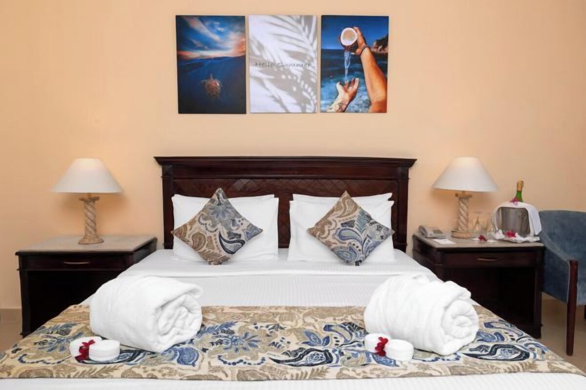 A hotel room with a bed, two nightstands, a lamp, a bottle, and artwork on the wall.