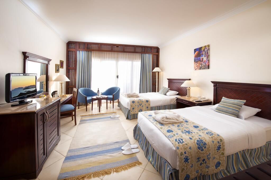 Een hotelkamer met twee bedden, een bureau met een televisie, een stoel, een klein tafeltje en een raam met gordijnen.