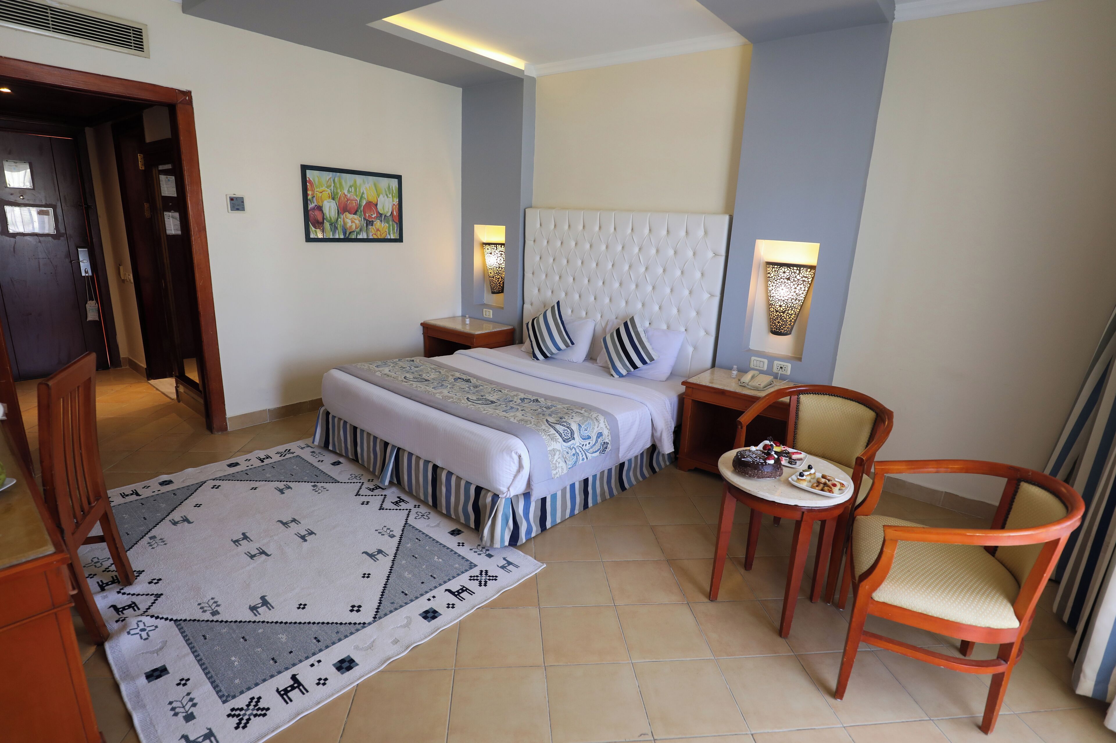 Una habitación de hotel con una cama grande, dos mesitas de noche, una pequeña mesa redonda con sillas, un tapete decorativo y un cuadro en la pared.