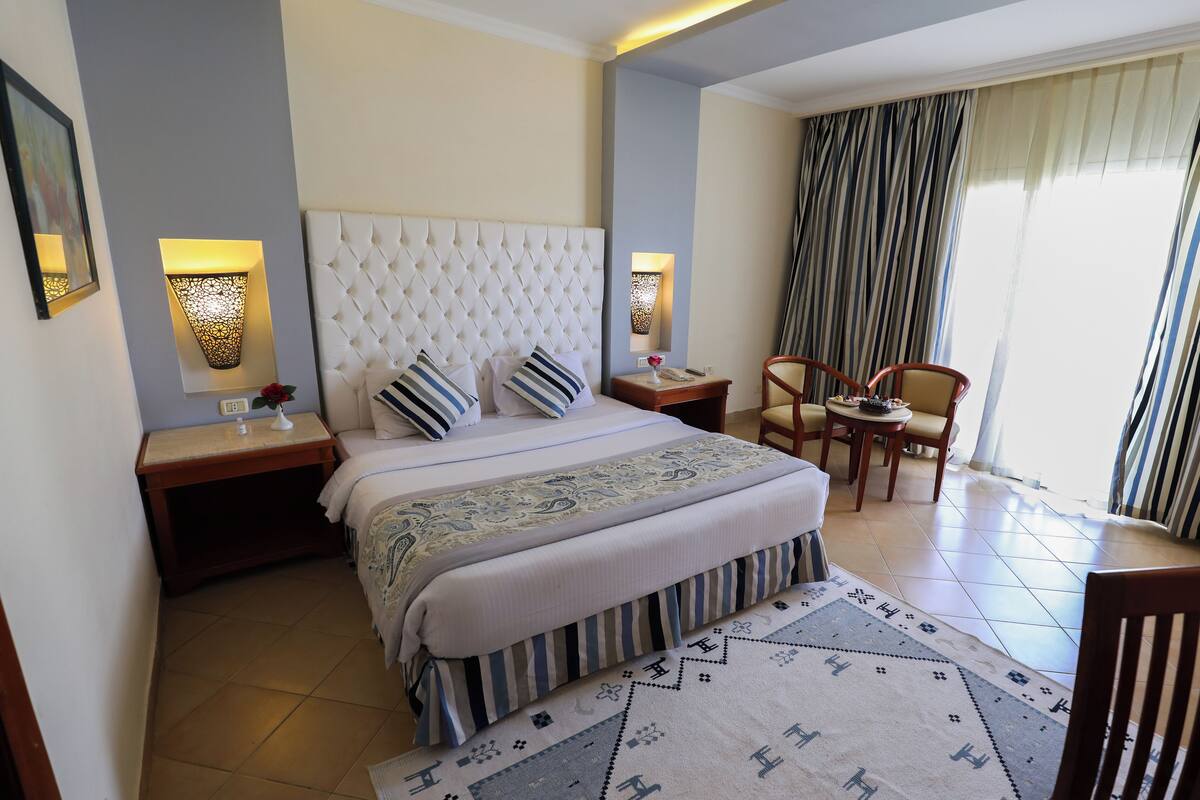 Ein Hotelzimmer mit einem großen Bett, zwei Nachttischen, einem Sessel, einem kleinen Tisch, einem gemusterten Teppich und Blick ins Freie durch Vorhänge.