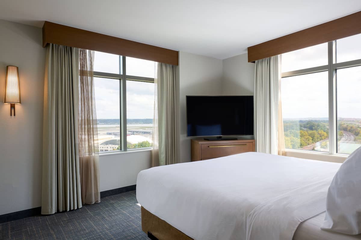 A hotel room with a large bed, a flat-screen TV, a wooden nightstand, and a view of a cityscape through large windows.