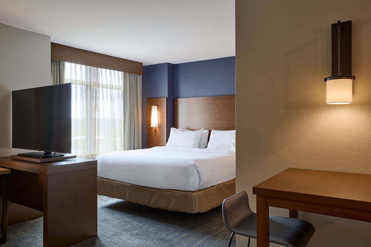 A hotel room with a bed, a desk with a television, a chair, and a wall-mounted light fixture.