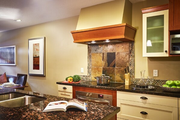 A kitchen with granite countertops, a stainless steel sink, a microwave, and a stove. There are cabinets, a backsplash, and a window with blinds. - Bellstar Suites at Solara Resort (Canmore)