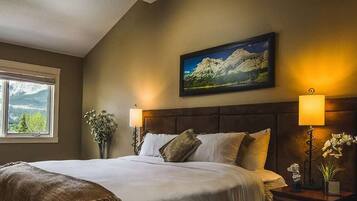 A bedroom with a large bed, two bedside lamps, a framed picture on the wall, and a window with a mountain view.