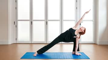 A person performing a yoga pose on a blue mat in a room with large windows and a wooden floor.