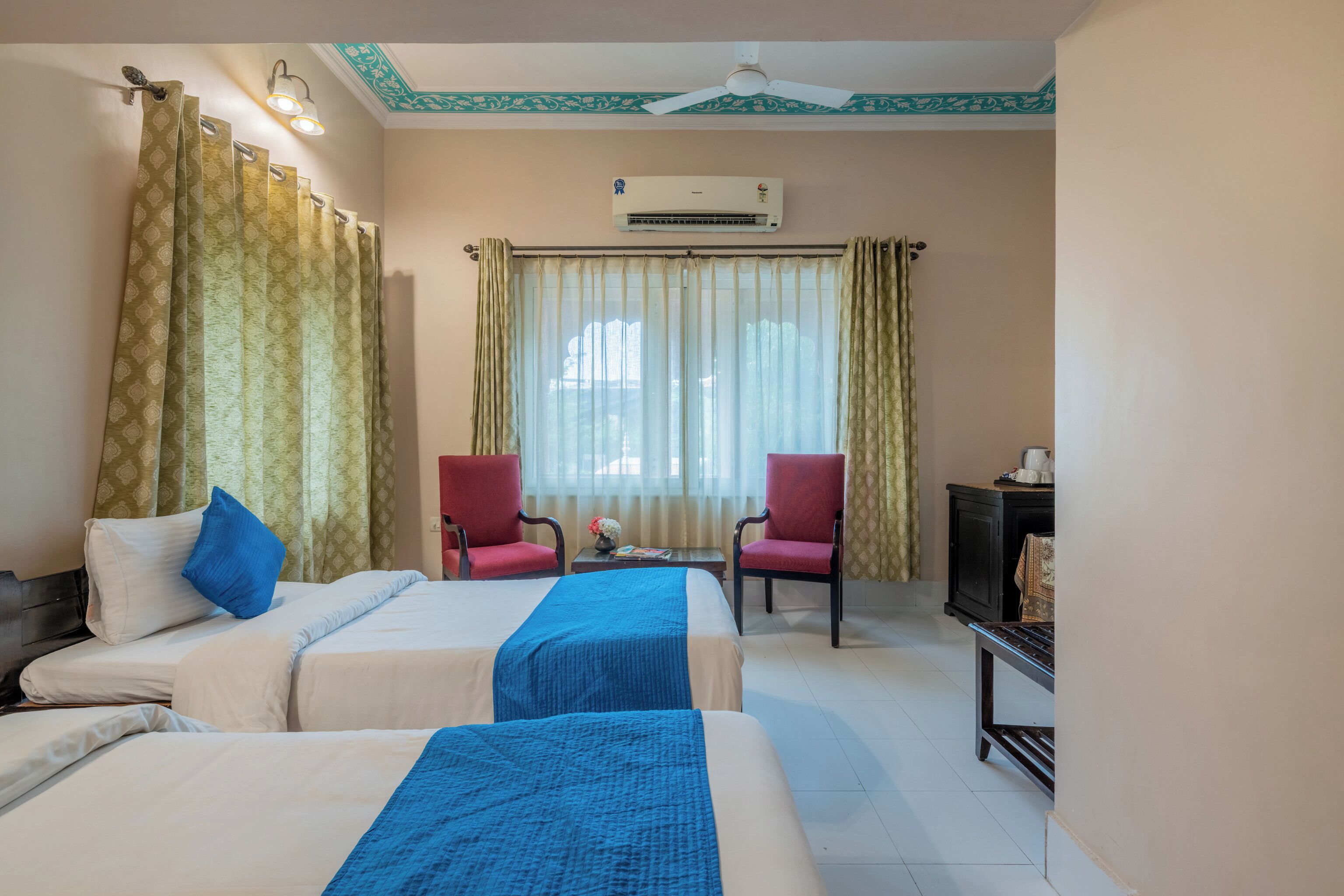 A hotel room with two beds, a ceiling fan, an air conditioner, and a small table with a lamp.
