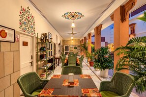 Un vestíbulo de hotel con una mesa de recepción, una estantería, sillas verdes, una mesa con diseño y un ventilador de techo.
