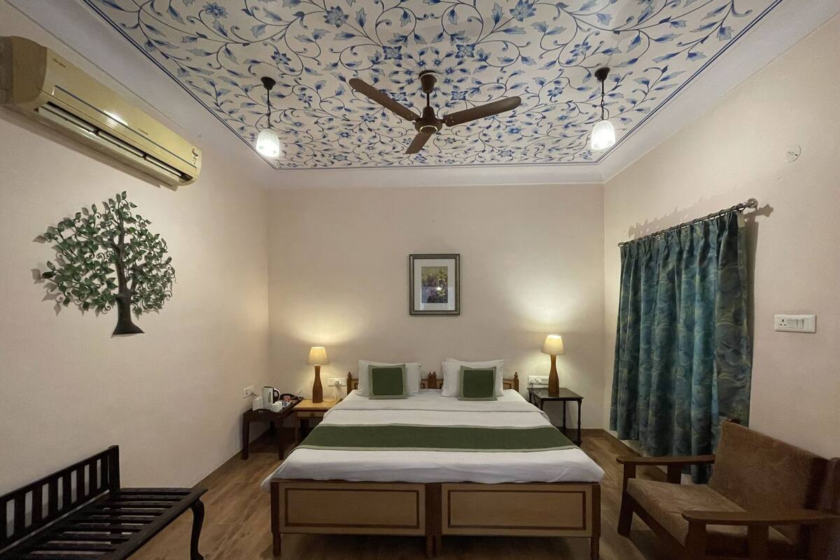 Ein Hotelzimmer mit einem Bett, zwei Nachttischen, einem Deckenventilator, einer Klimaanlage, einer Wanddekoration mit Baummotiv, einem gerahmten Bild und einem Fenster mit Vorhängen.