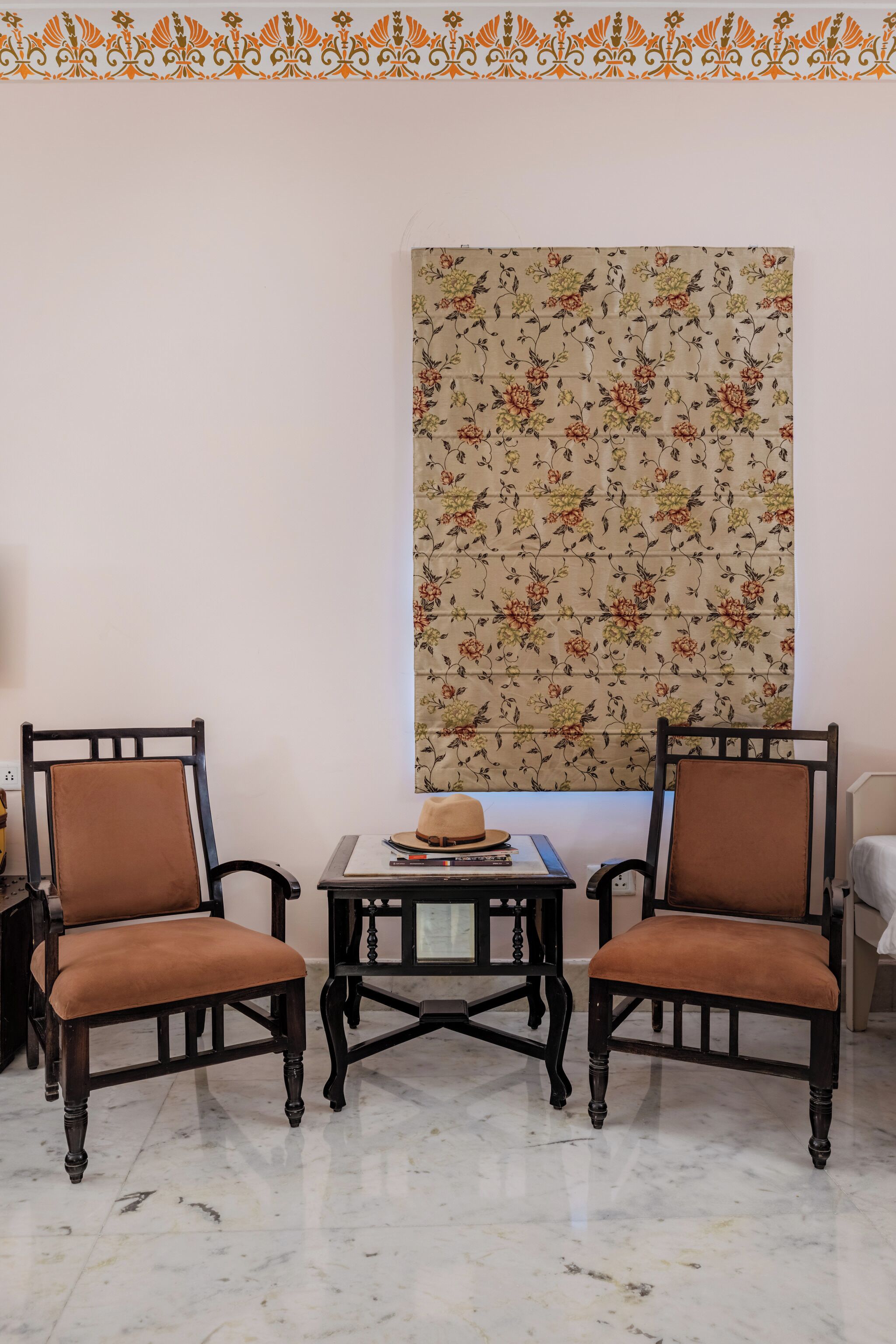 Dos sillones marrones con patas negras, una mesita con un sombrero y libros, y un tapiz con estampado floral en la pared.